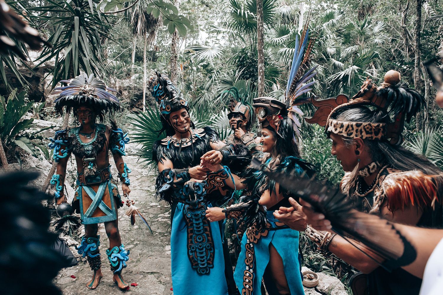 Group of performers in elaborate Aztec-Mayan ceremonial costumes interact in a tropical jungle setting.