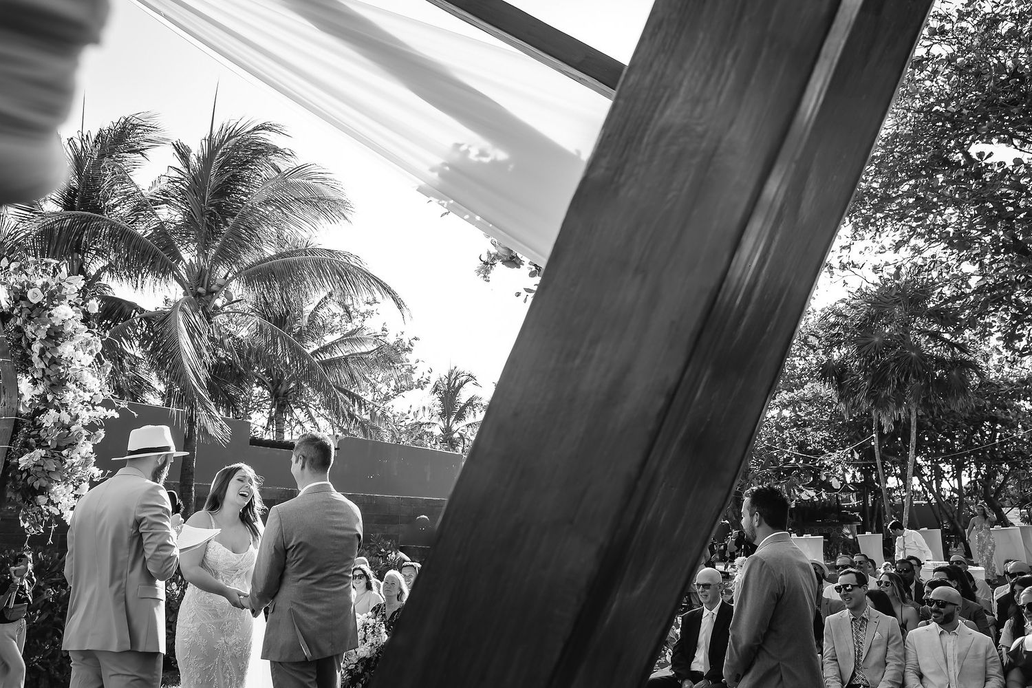 Black and white photo of wedding guests gathered outdoors near a modern sculpture with tropical palm trees in background.