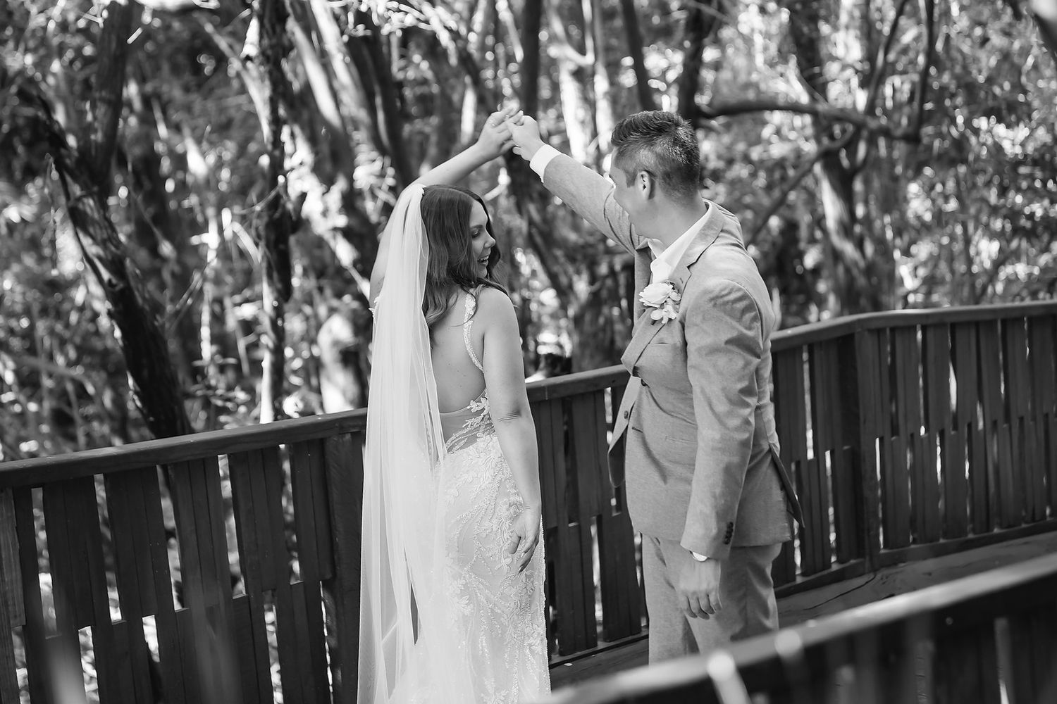 Black and white photo of a groom spinning his bride in a wedding dress on a wooden deck surrounded by trees.