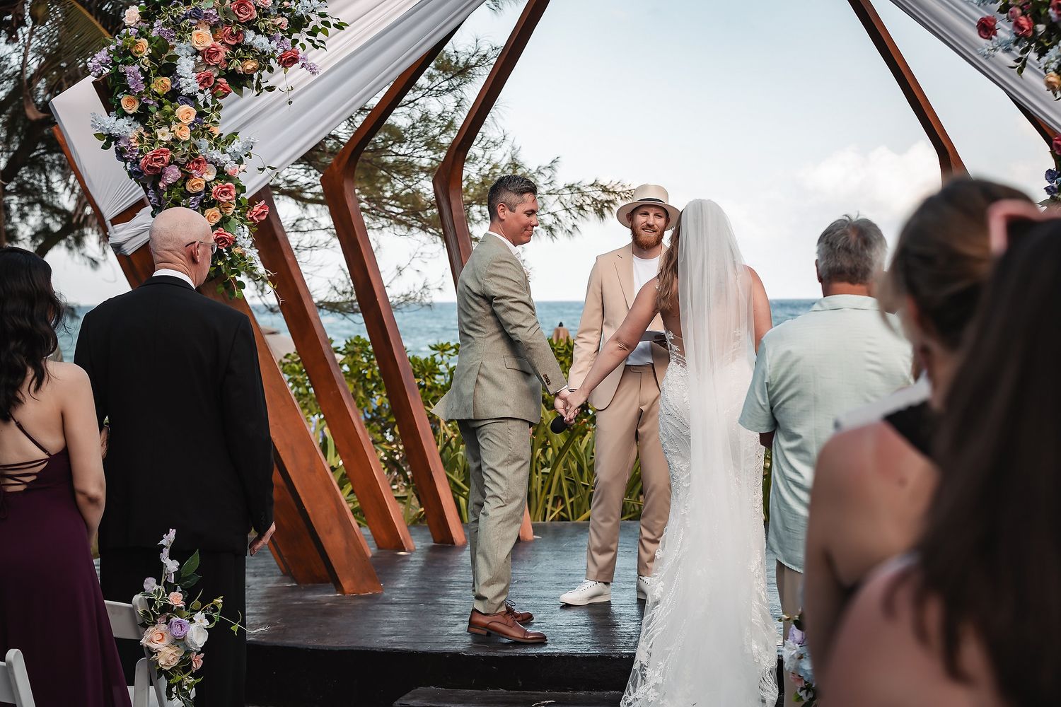 Couple exchanges vows at outdoor beach wedding ceremony under floral arch with ocean view.