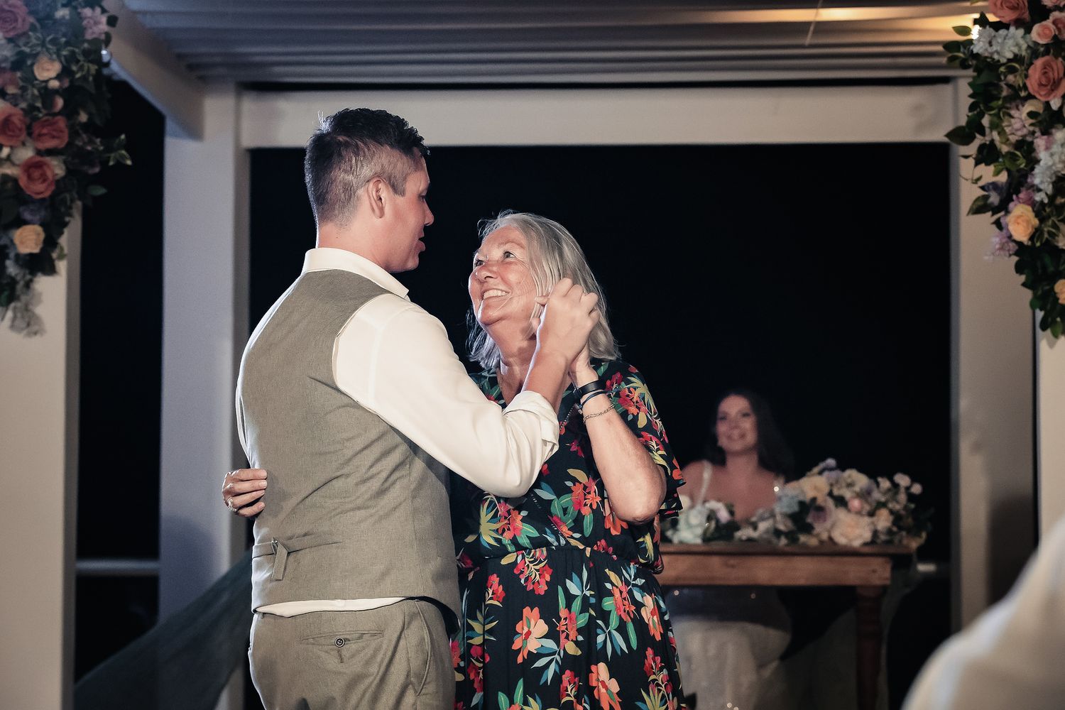 Groom sharing a tender mother-son dance at wedding reception, with floral arrangements and guests visible in background.