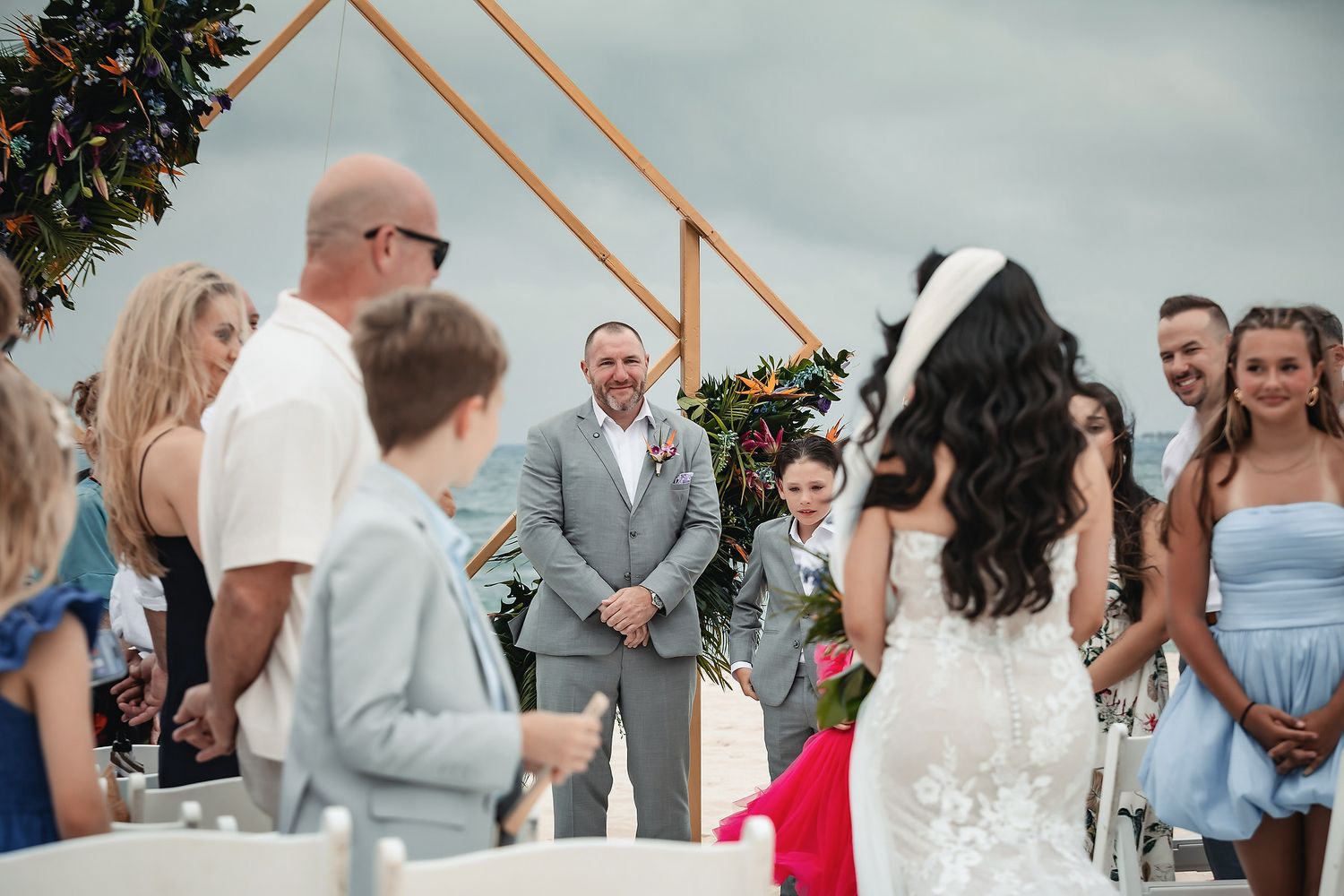 Groom smiling at bride during outdoor beach wedding ceremony with geometric copper arch and guests seated.