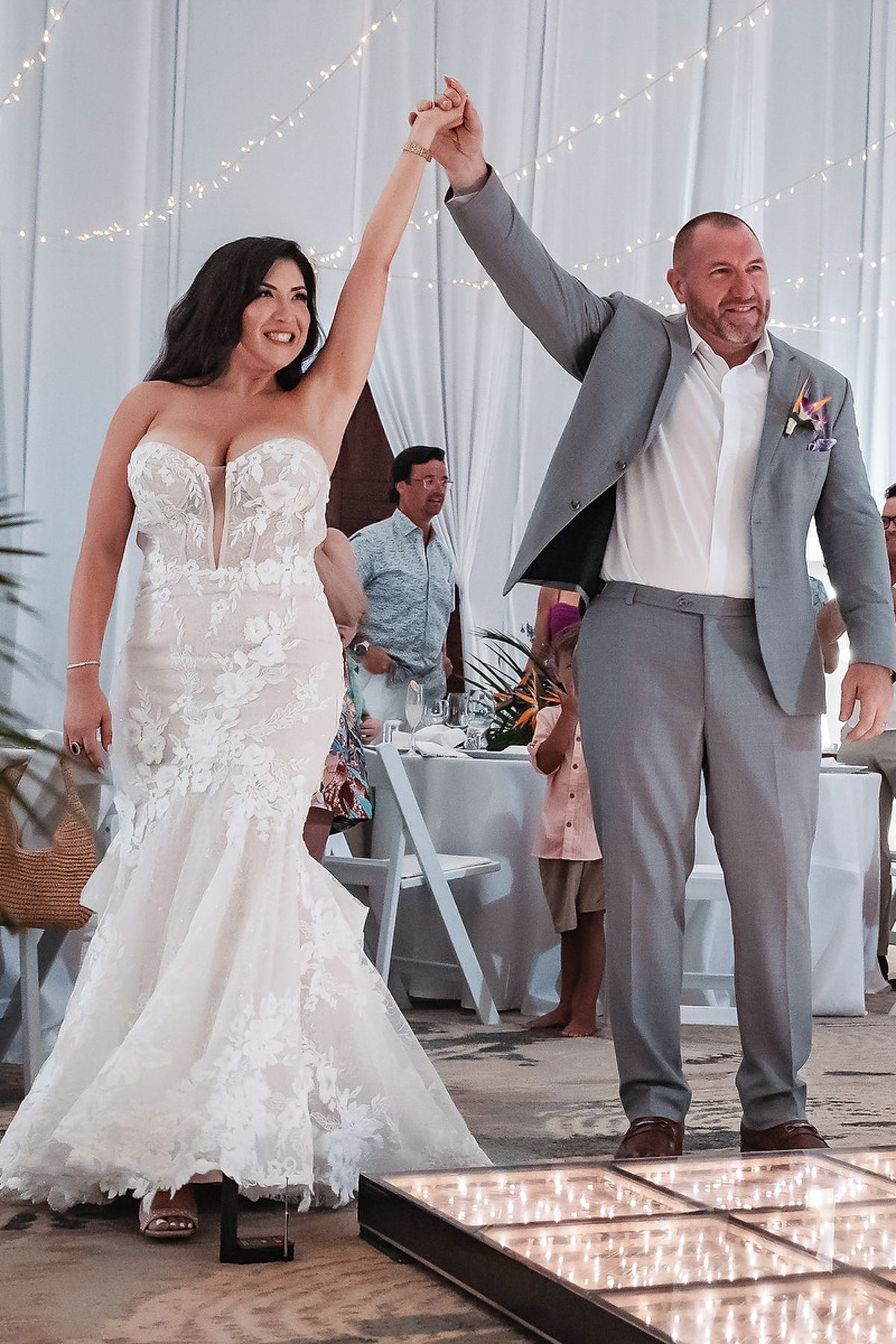 Bride in lace mermaid gown and groom in gray suit raise hands triumphantly on illuminated dance floor at wedding reception.