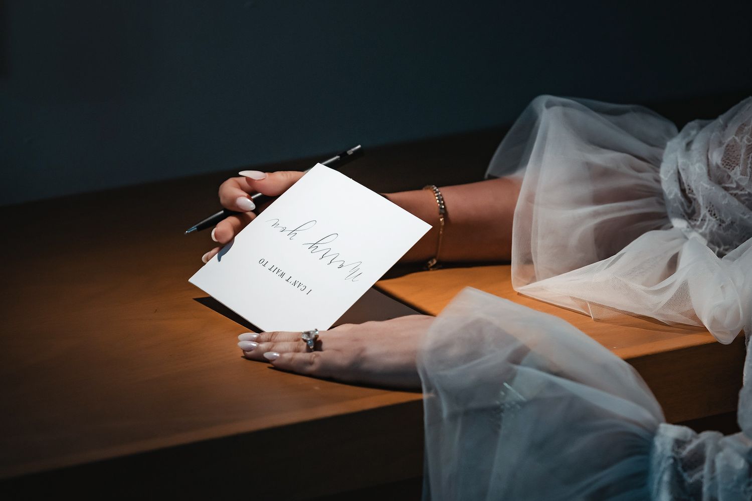 Bride in white tulle dress signing wedding guest book with pen at wooden table, wearing elegant ring.