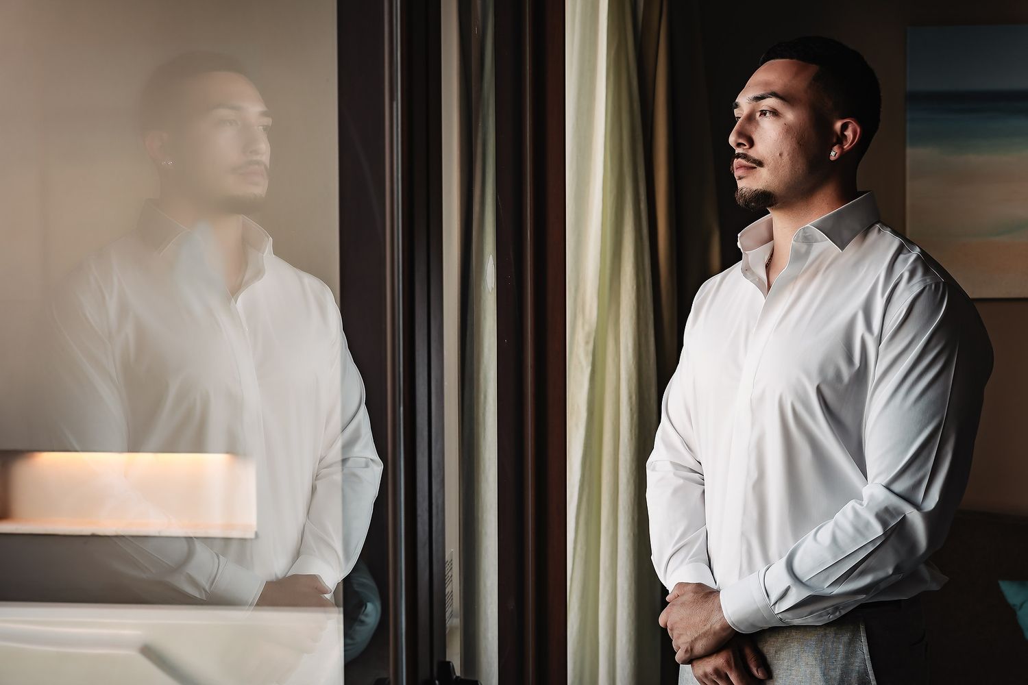Man in white dress shirt gazing thoughtfully out a window, with his reflection visible in the glass.
