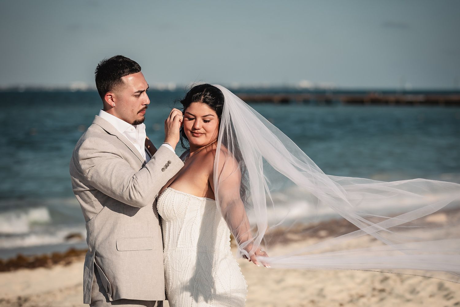 Groom in tan suit adjusting brides veil on a sunny beach wedding with ocean backdrop.