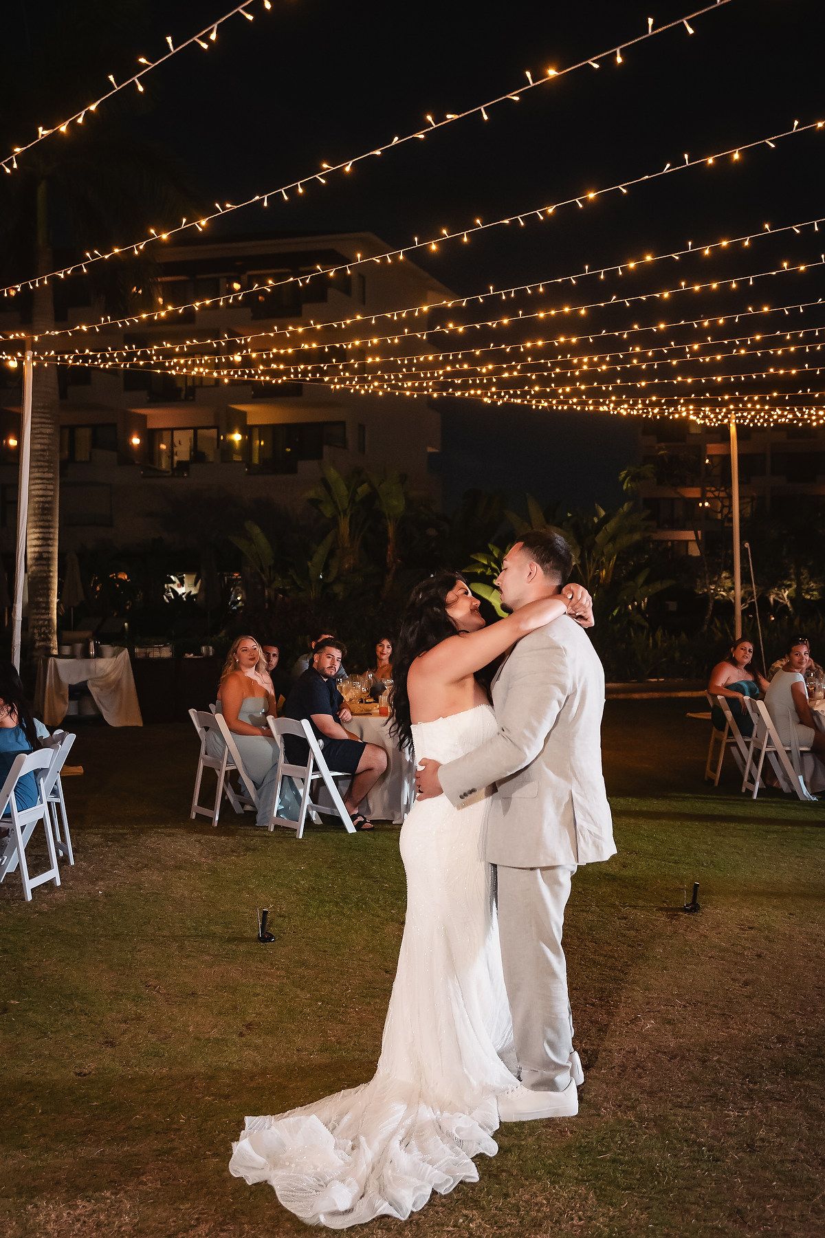 Bride and groom share their first dance under romantic string lights at an outdoor evening wedding reception.