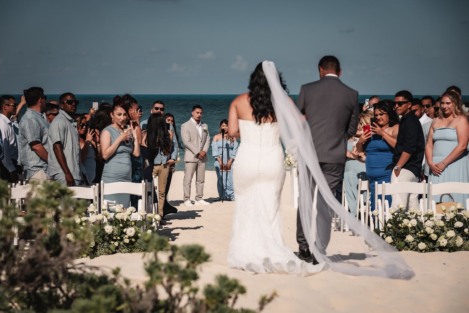 Bride and groom walk down the aisle at a romantic beach wedding ceremony with guests seated on white chairs.