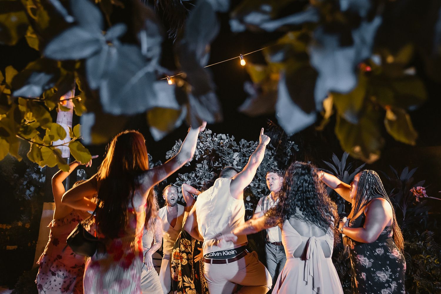 Bridal party dancing outdoors at night under string lights and lush trees during a wedding celebration.
