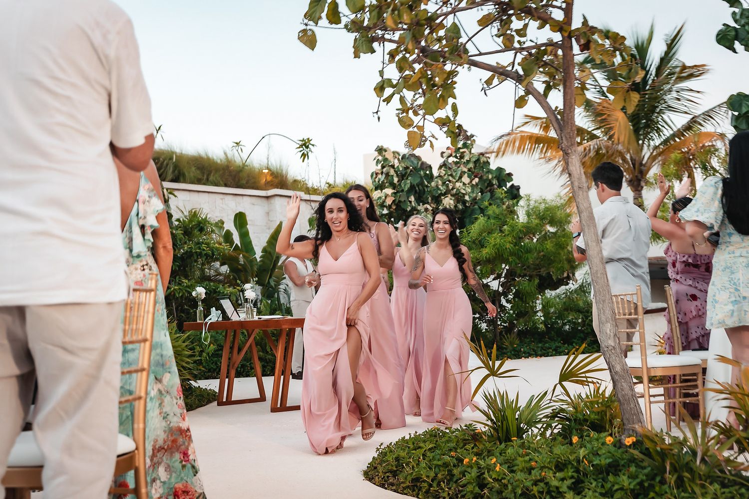 Bridesmaids in pink dresses walking down outdoor aisle at tropical beach wedding ceremony.