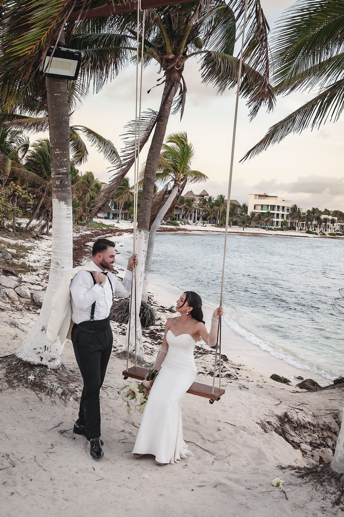 Bride sits on beach swing by palm tree as groom stands beside her, ocean backdrop at their tropical wedding.