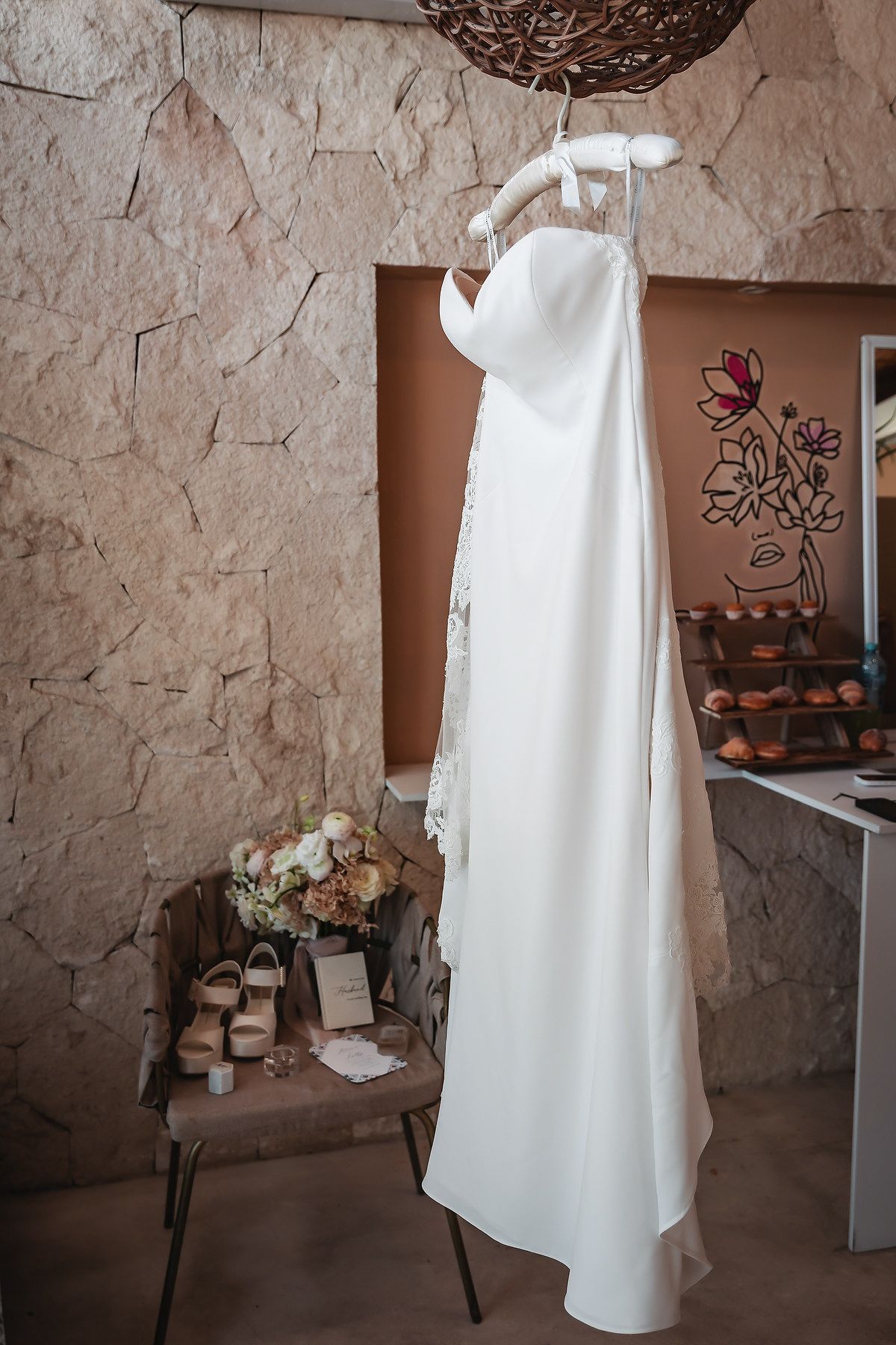 Elegant white strapless wedding dress hanging on a decorative hook against a stone wall, with bridal shoes and bouquet below.