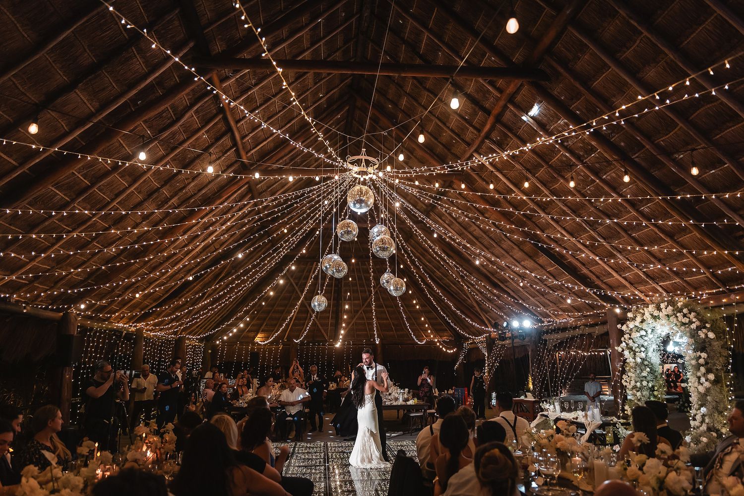 Romantic barn wedding reception with fairy lights draped across wooden ceiling, couple dancing surrounded by elegant floral arrangements.