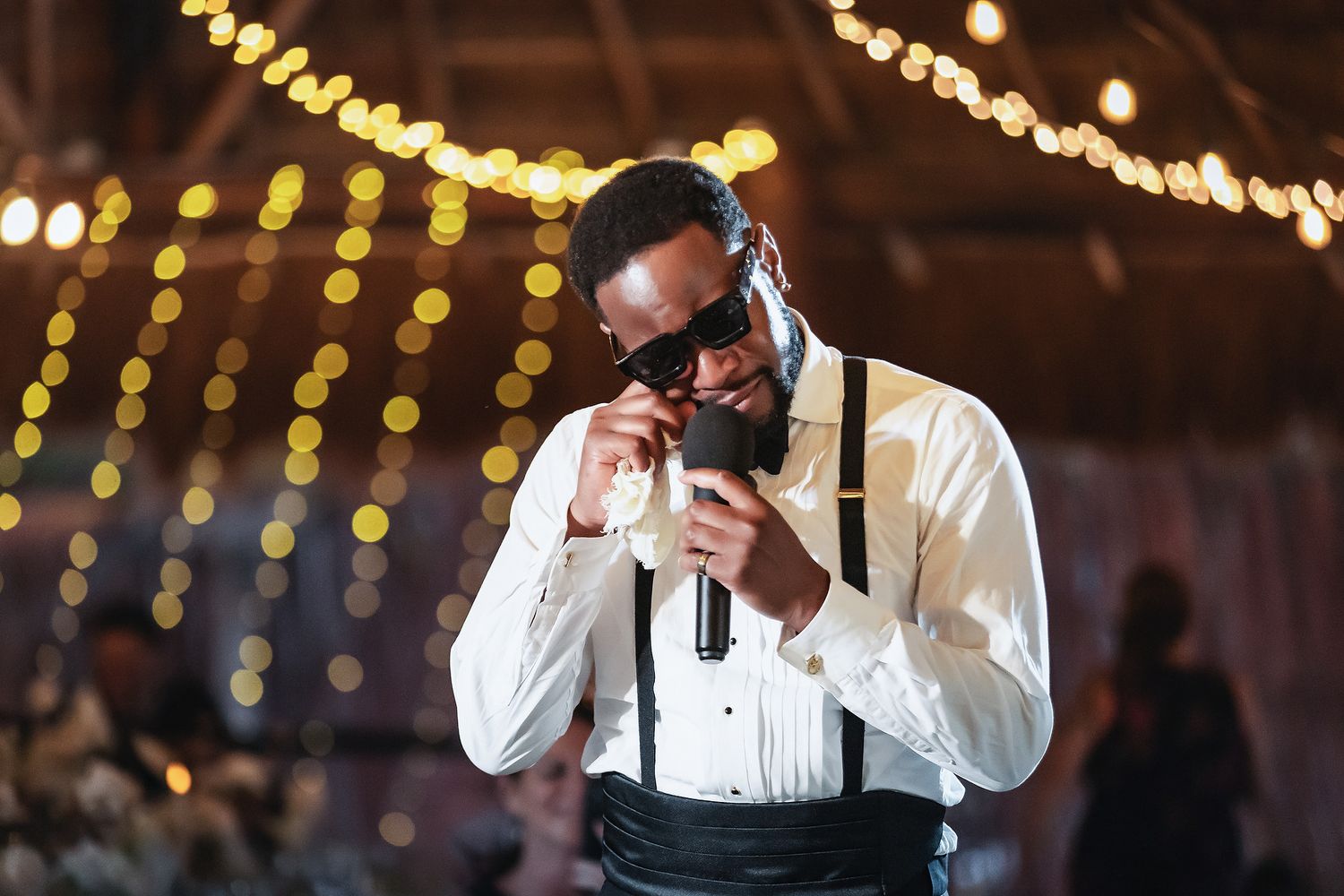 Groom in white shirt and suspenders adjusting bow tie at elegant wedding reception with bokeh lights.
