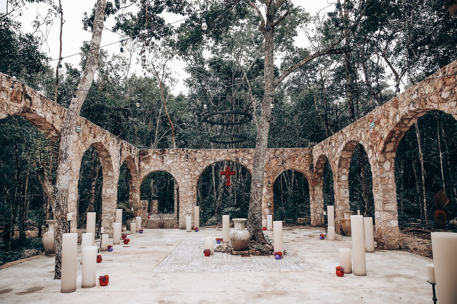 Stone colonnade with multiple arches in a forest setting decorated with red candles and white pillars.