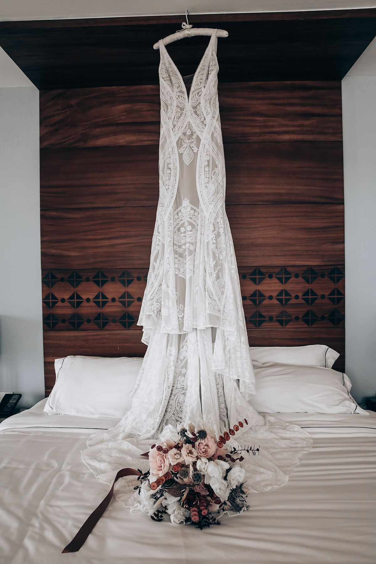 White lace wedding dress hanging above a bed with a pink and white floral bouquet displayed on white bedding.