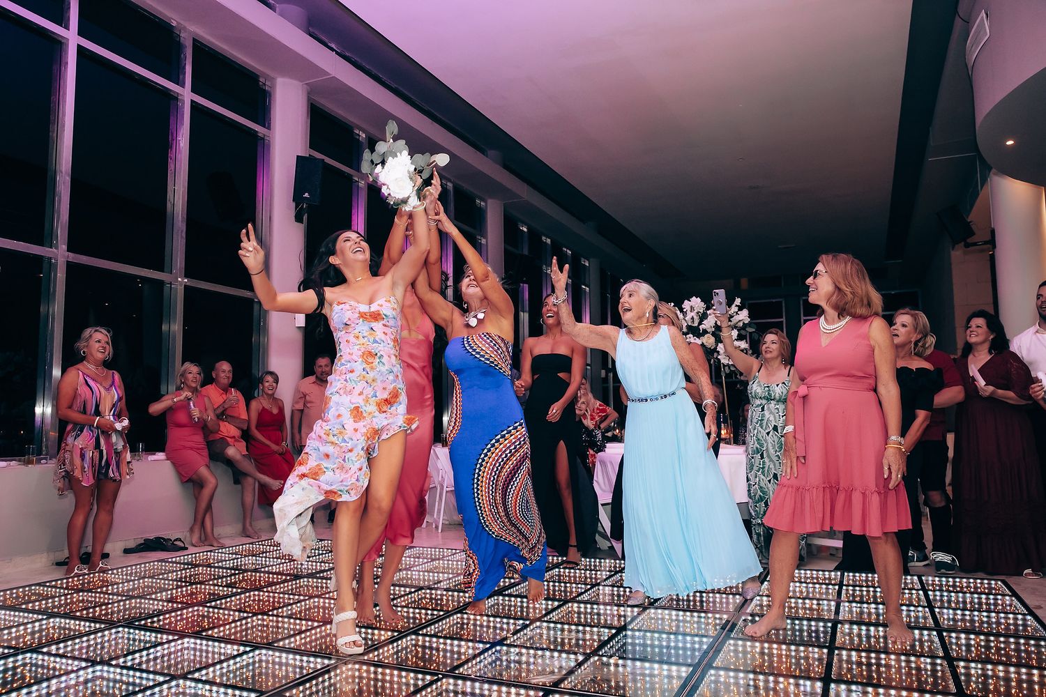 Wedding reception dance party with guests on an illuminated floor under pink ambient lighting.