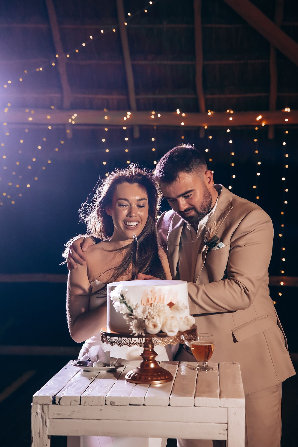 A couple in formal attire cuts their wedding cake together in a dimly lit venue decorated with string lights.