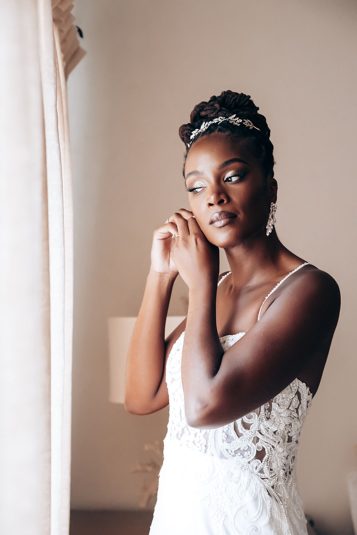 Bride in white lace dress putting on earrings in natural window light.