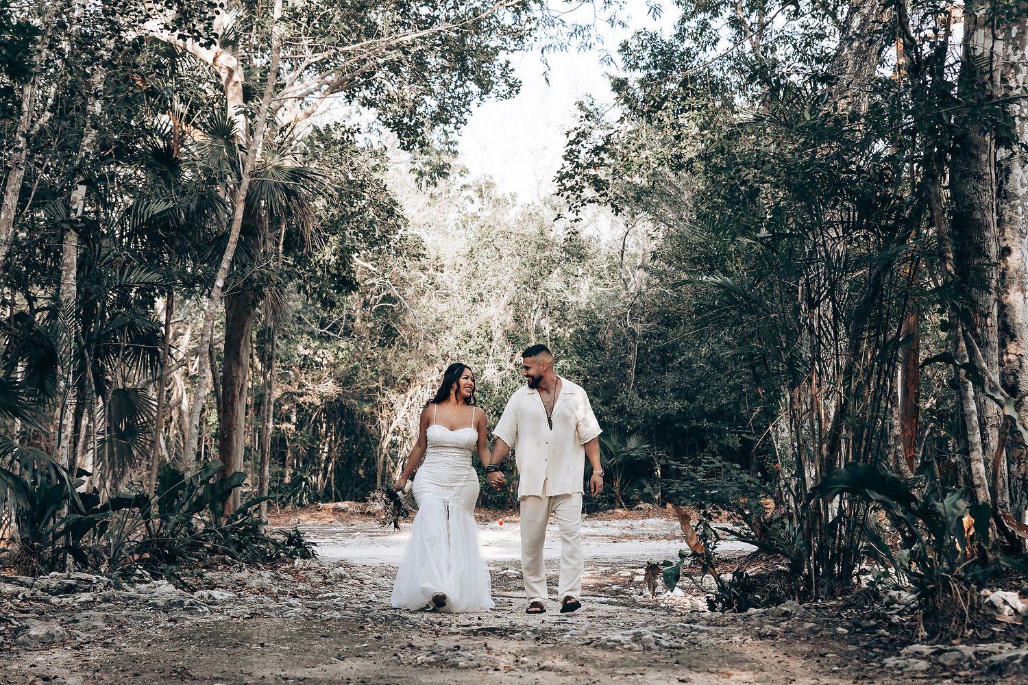 A couple walks hand in hand through a forest path in wedding attire.