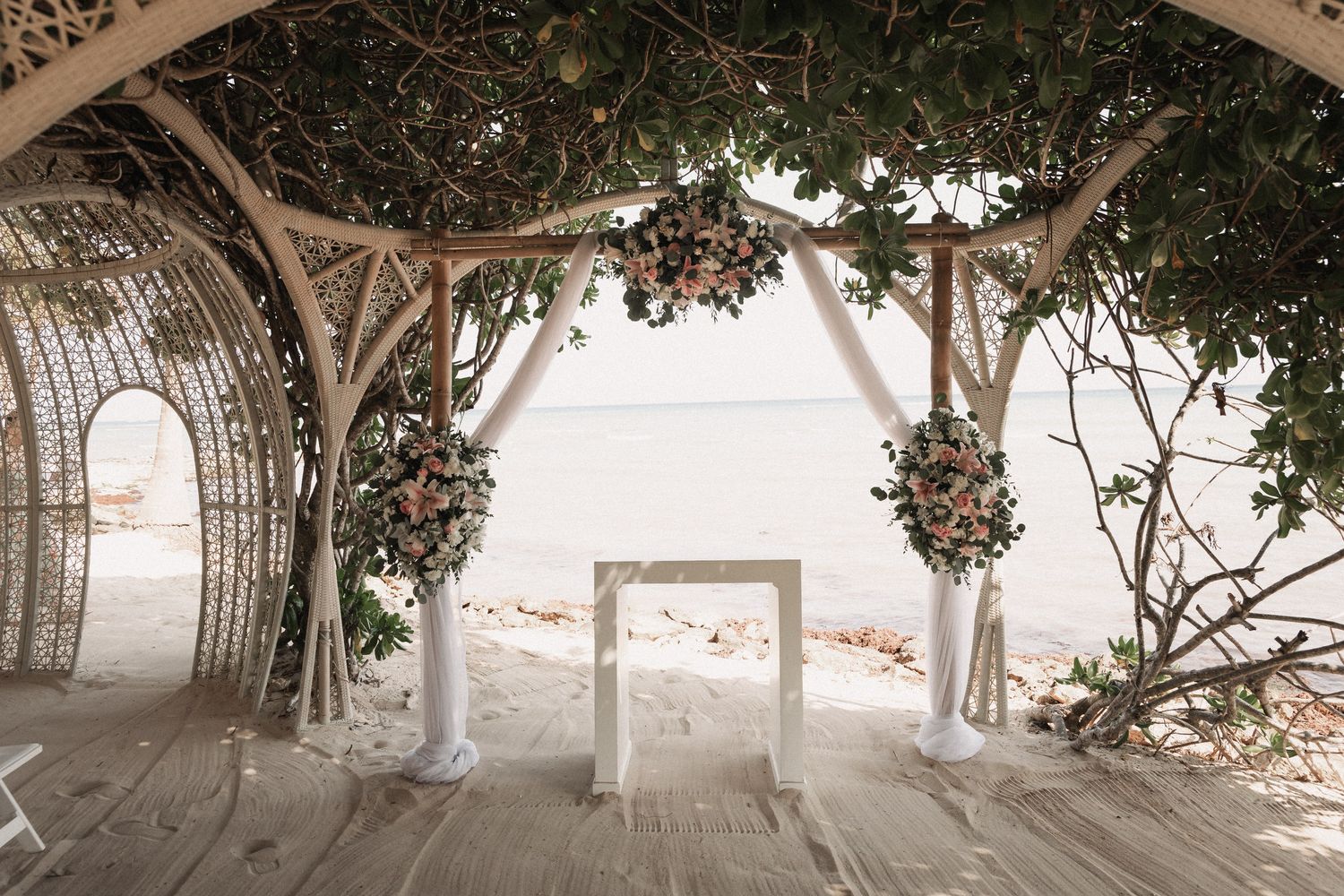 Romantic beach wedding ceremony setup with floral arch, white altar, and ocean view under shade structure.