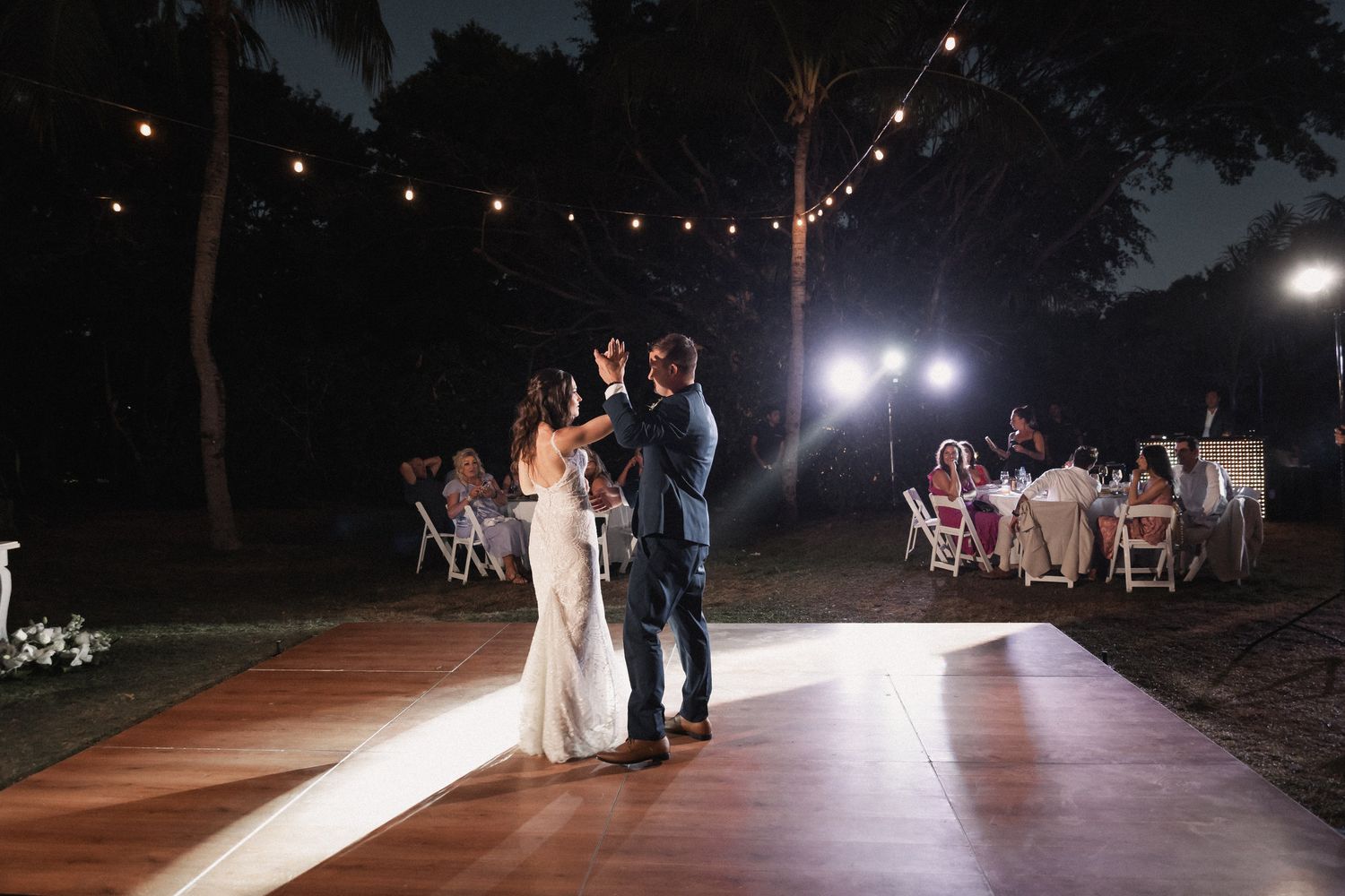 Bride and groom share first dance on outdoor wooden floor under string lights at night.