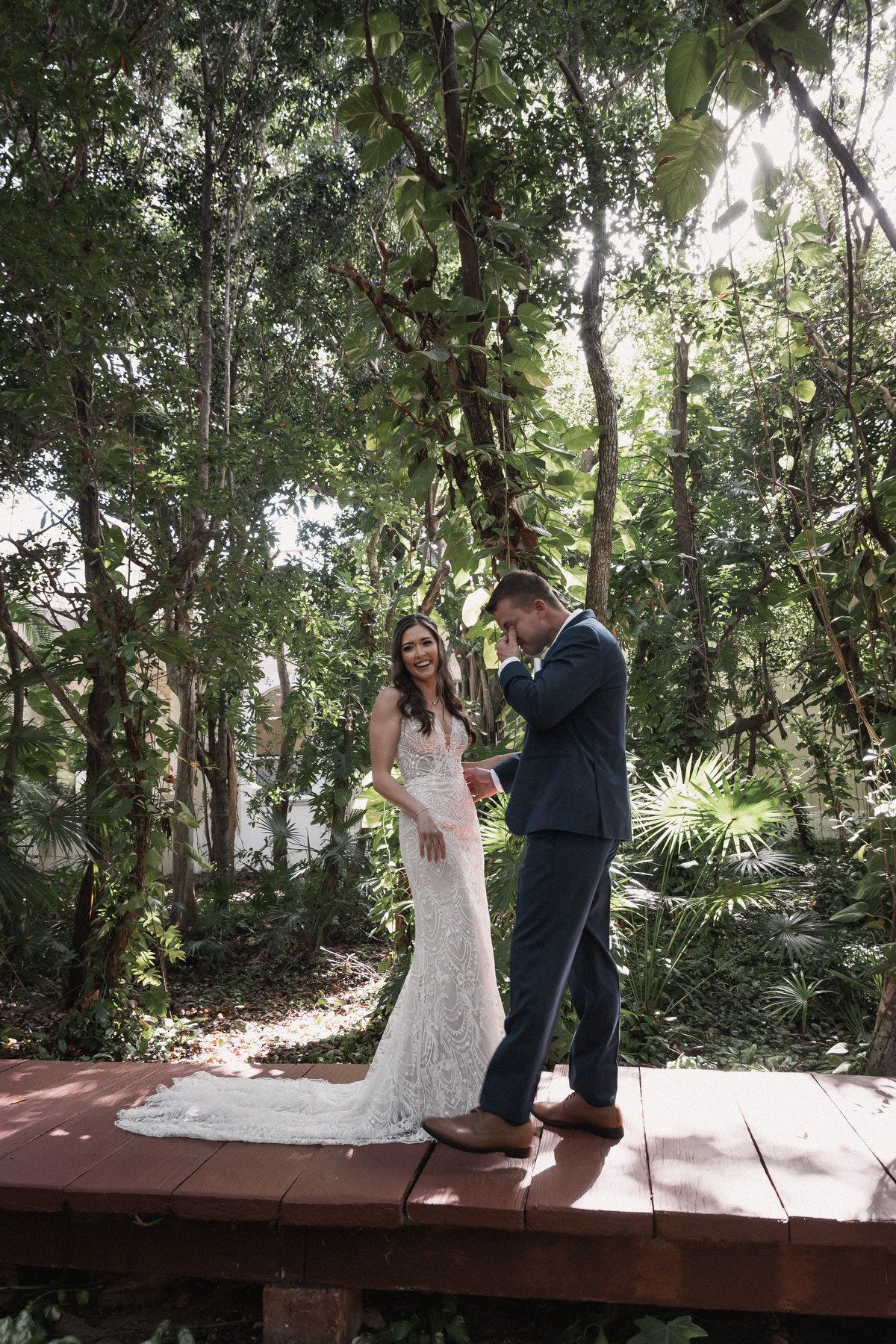 Bride and groom laugh together during first look on wooden deck in sunlit tropical garden setting.