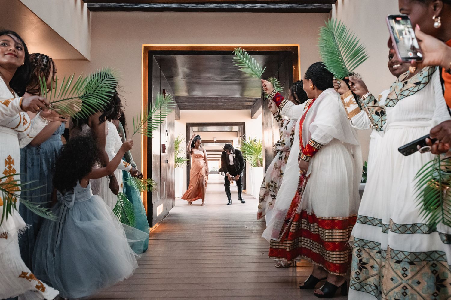Guests waving palm leaves welcoming couple in traditional Ethiopian wedding attire down decorated hallway.