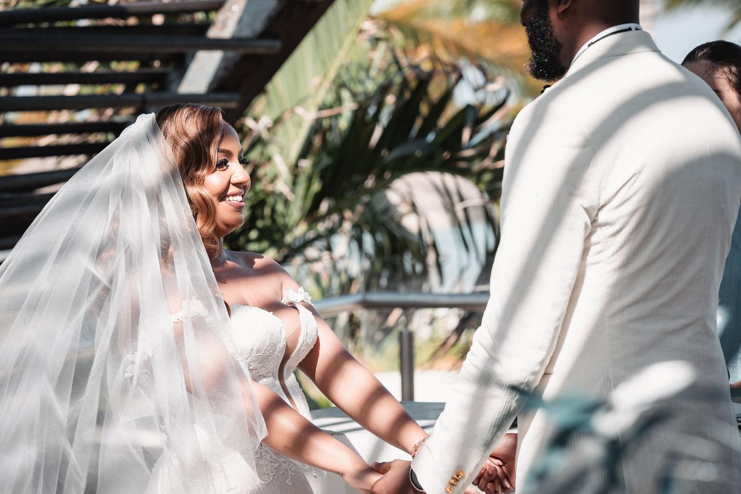 Bride smiling joyfully at groom while holding hands during outdoor wedding ceremony with veil.