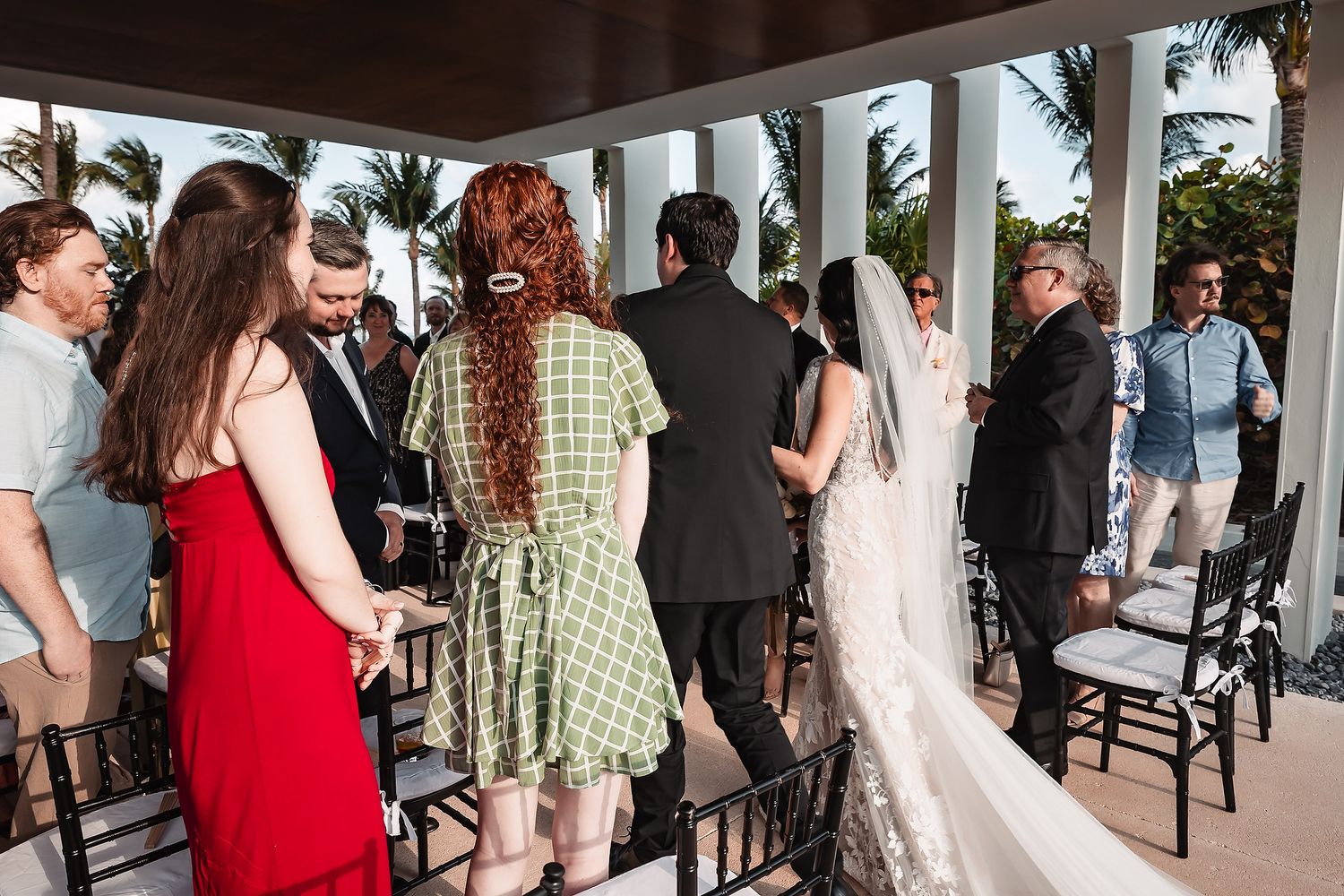 Guests gather at an outdoor wedding ceremony on a tropical venue with white columns and palm trees.