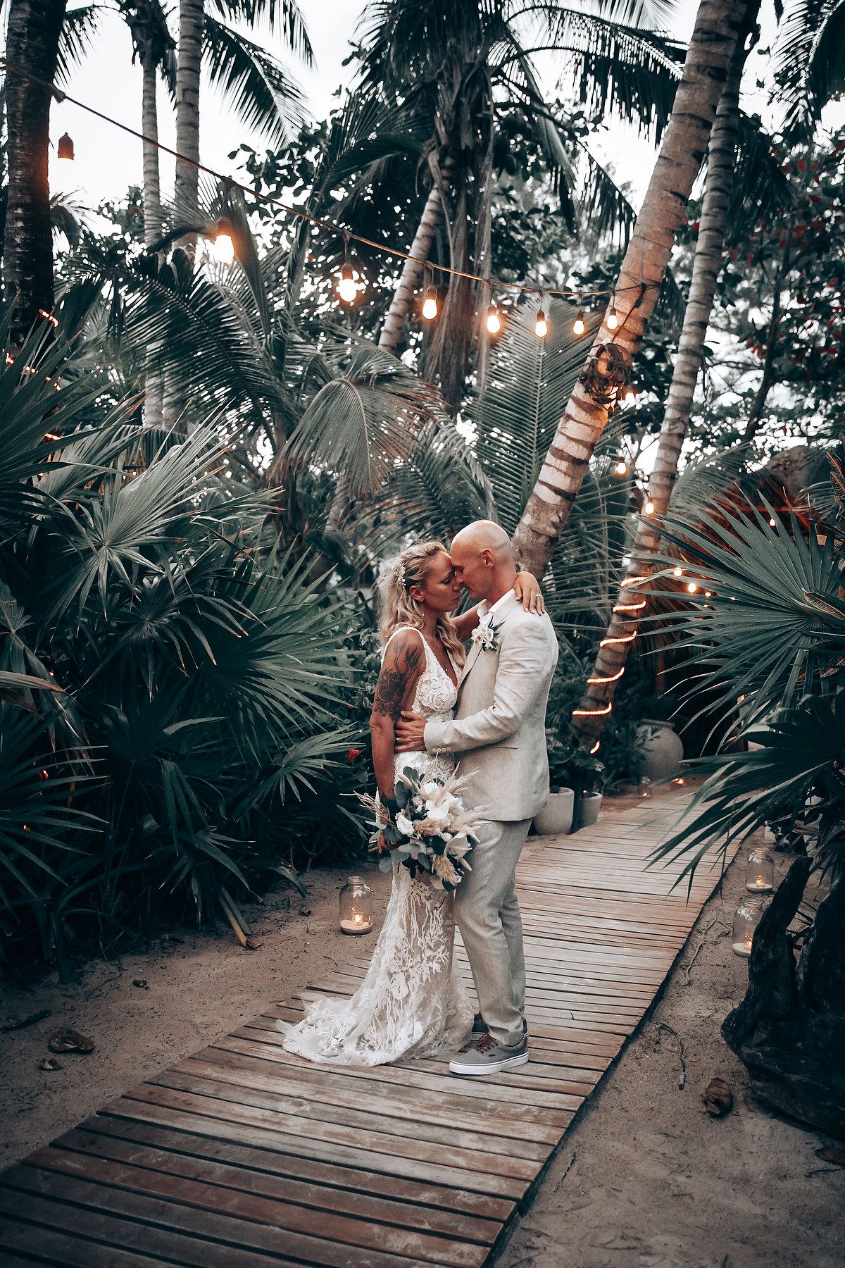 Newlyweds share an intimate moment on a wooden walkway surrounded by tropical palm trees at dusk.
