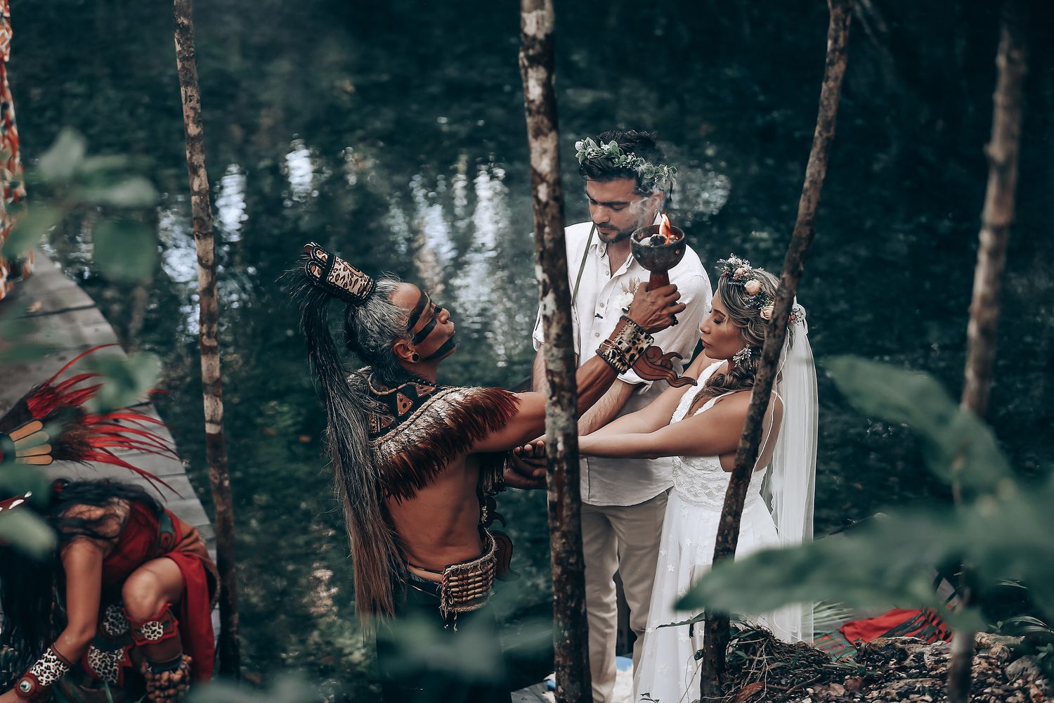 A couple in white attire embraces in a romantic woodland setting beside a tranquil pond.