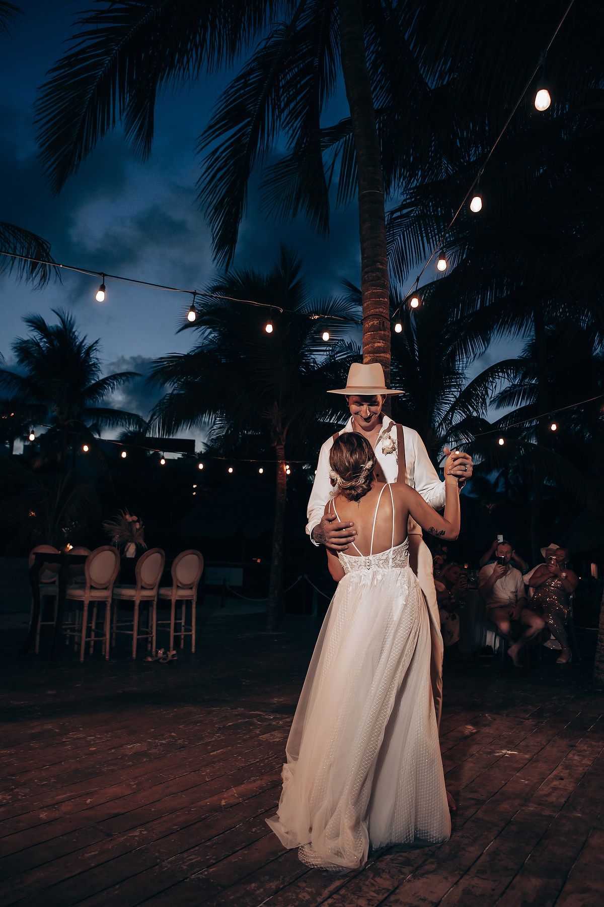 A romantic first dance under string lights and palm trees at a beach wedding reception.