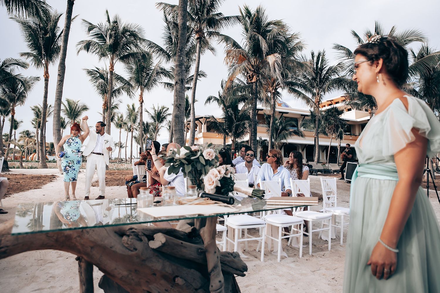 Beachfront wedding reception setup with white tables and chairs surrounded by swaying palm trees at sunset.
