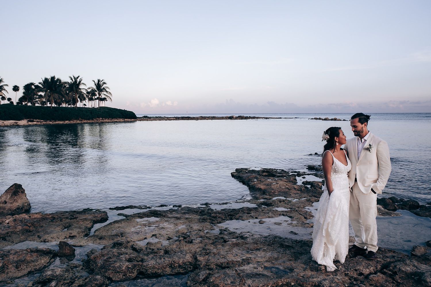 A romantic beach wedding scene at sunset with a couple standing on rocky shoreline.