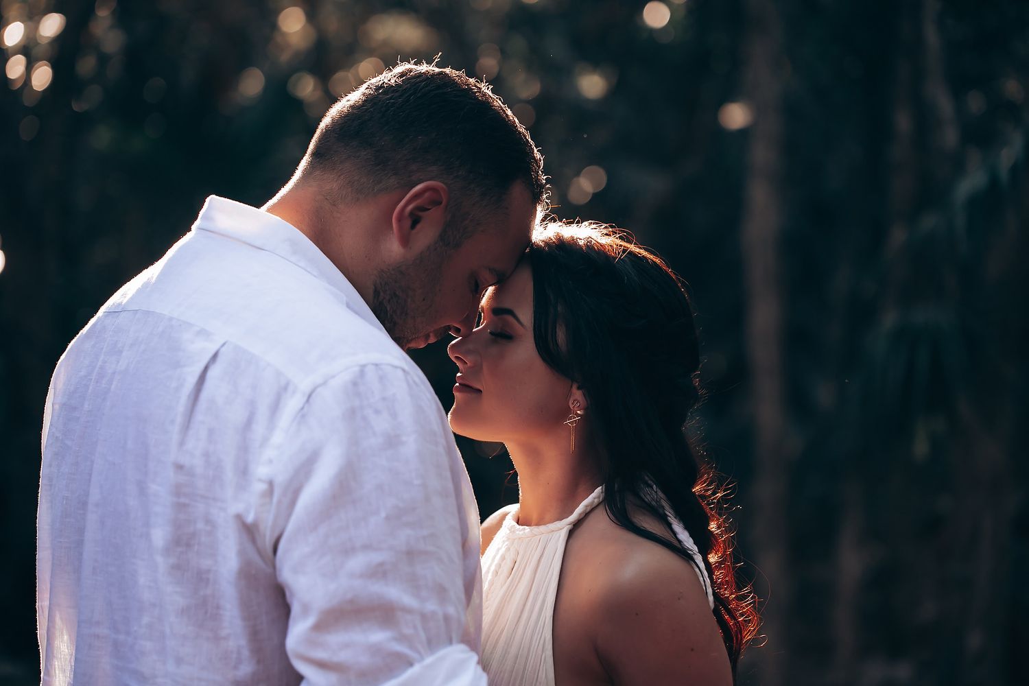 A couple shares a romantic moment in soft evening lighting with a dark forest backdrop.