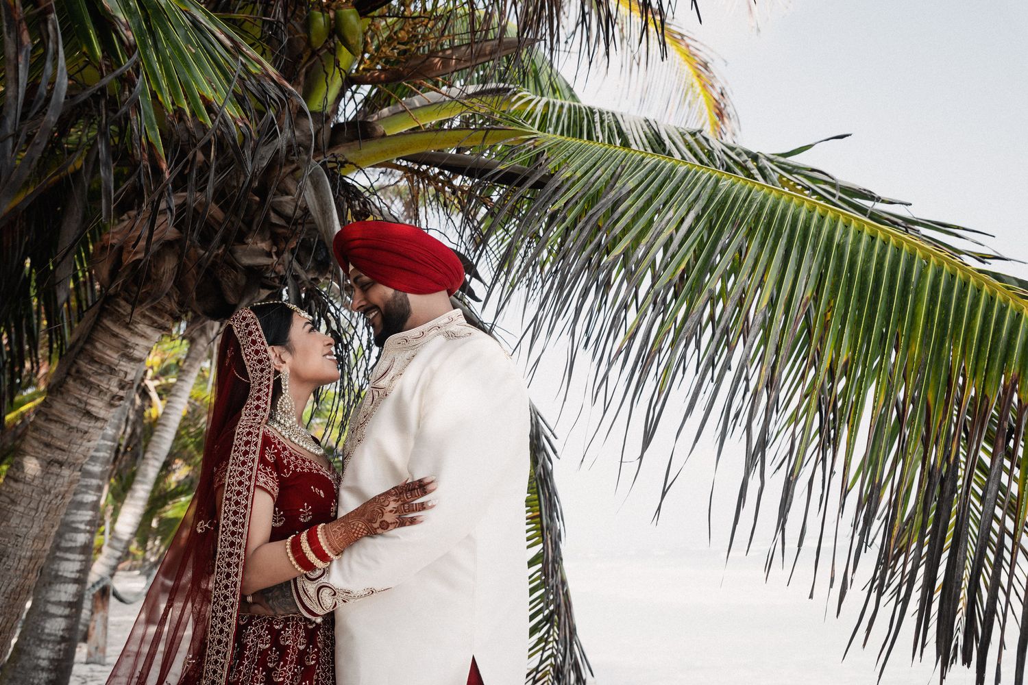 Sikh bride in red lehenga and groom in white sherwani pose romantically under tropical palm trees.