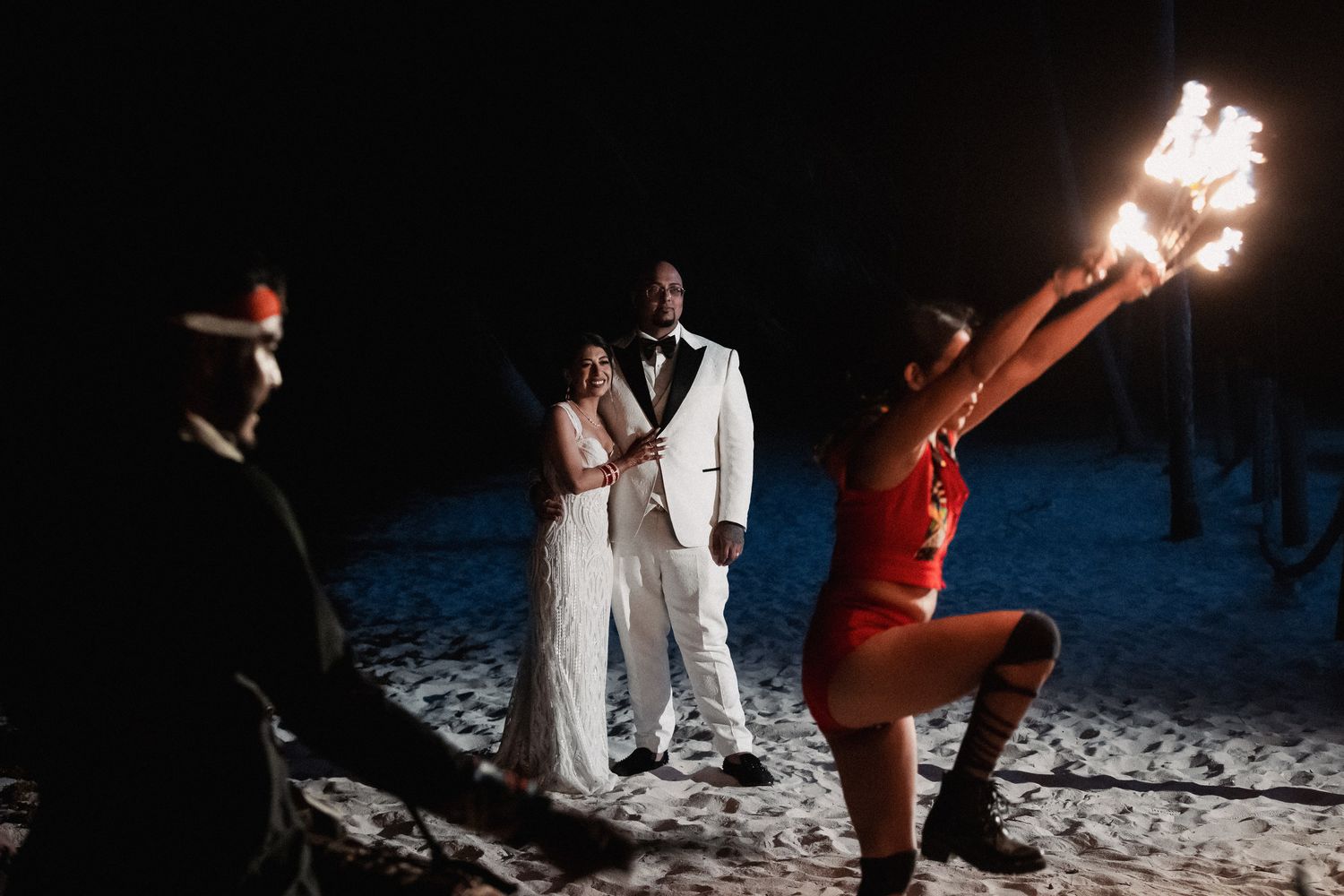 Bride and groom watch a fire dancer perform on the beach during their romantic outdoor nighttime wedding celebration.