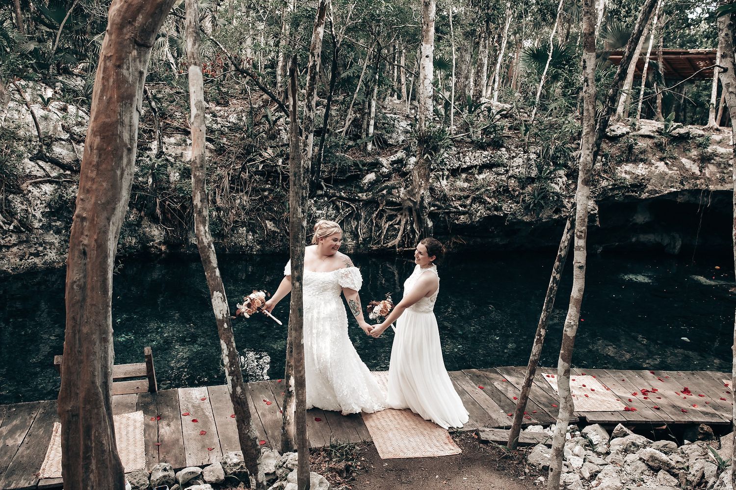 Two brides in white wedding dresses hold hands in a lush, outdoor setting.