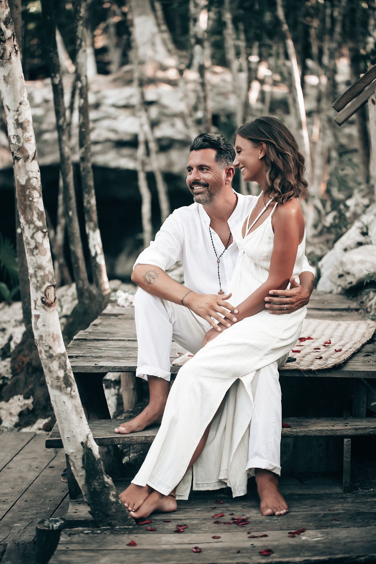 A couple dressed in white clothing sitting on a wooden bridge in a forest setting.