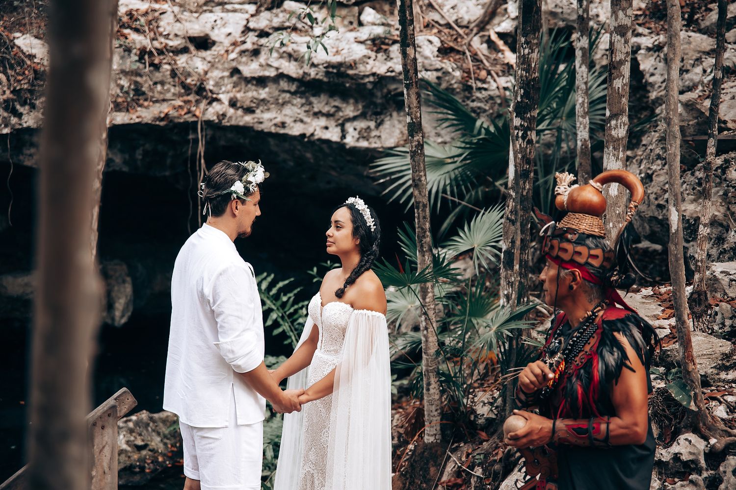 A couple in white outfits poses together in a lush tropical garden setting for wedding photos.