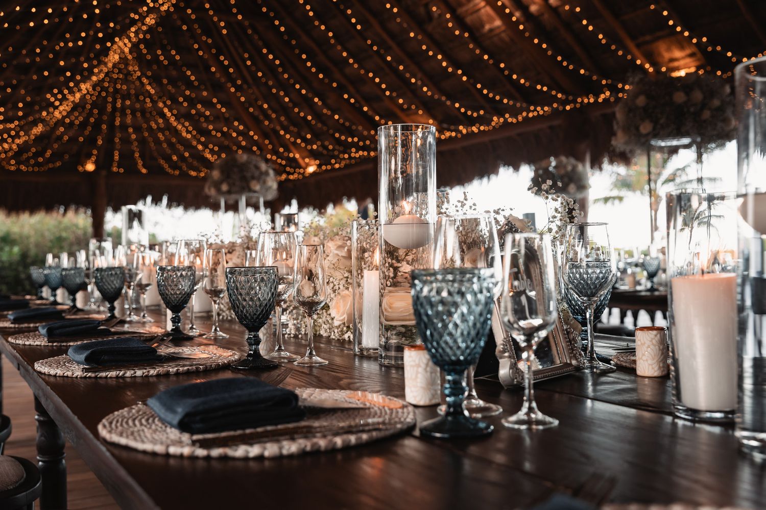 Romantic outdoor wedding reception table with blue glassware, candles, and string lights overhead.