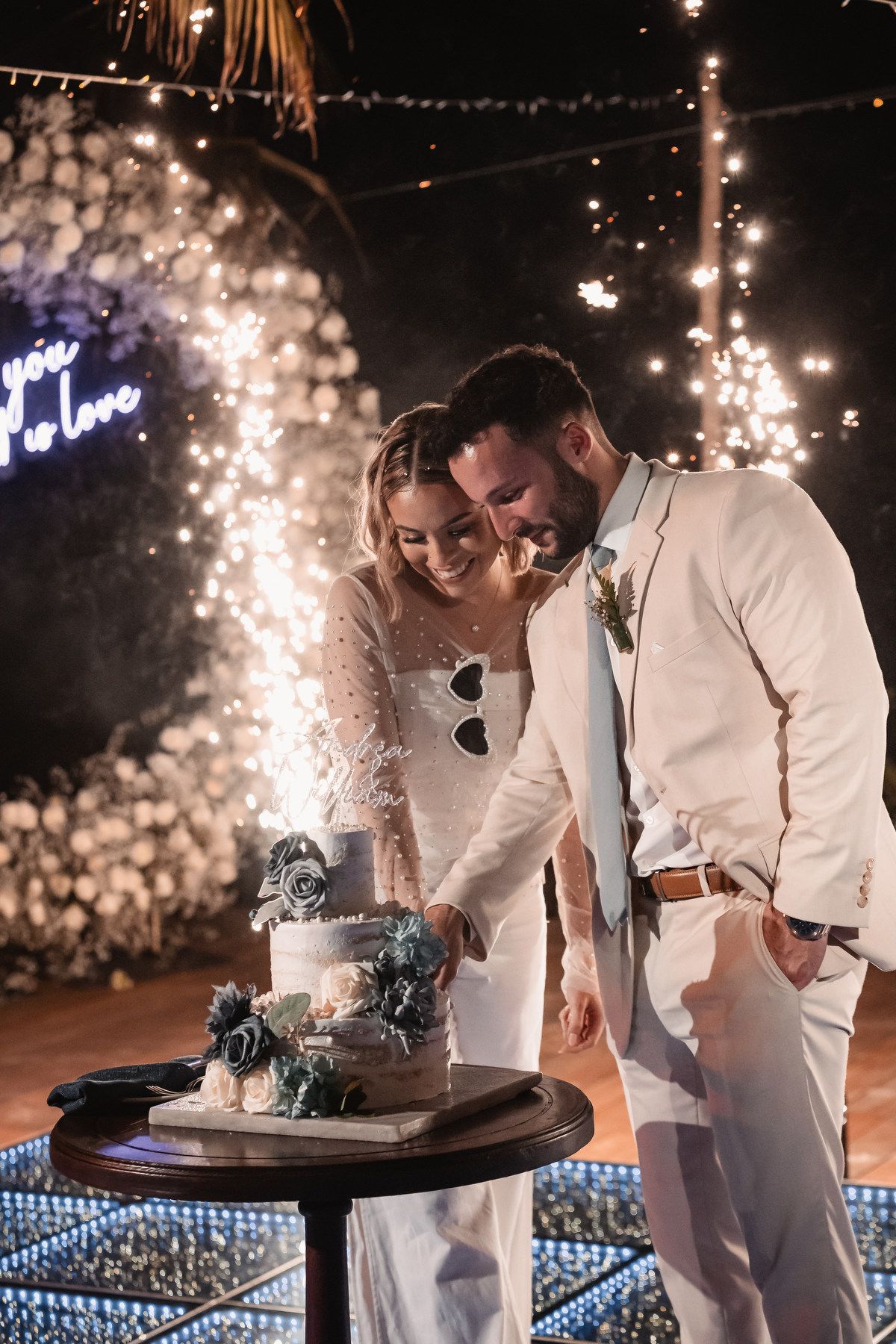 Smiling newlyweds cutting tiered wedding cake surrounded by sparkling fireworks at night.