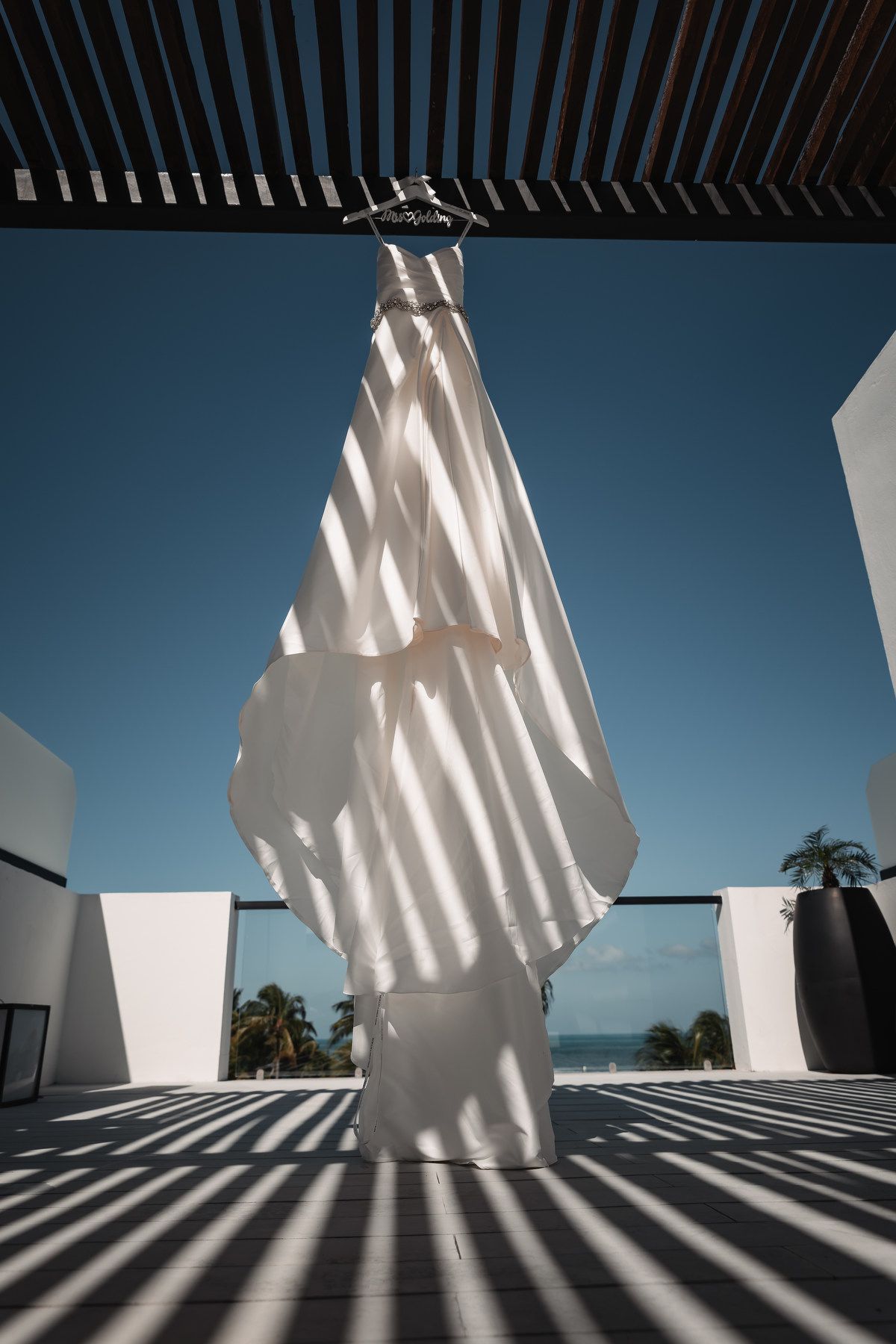 White wedding dress hanging on pergola with ocean view and striped shadow pattern on ground.