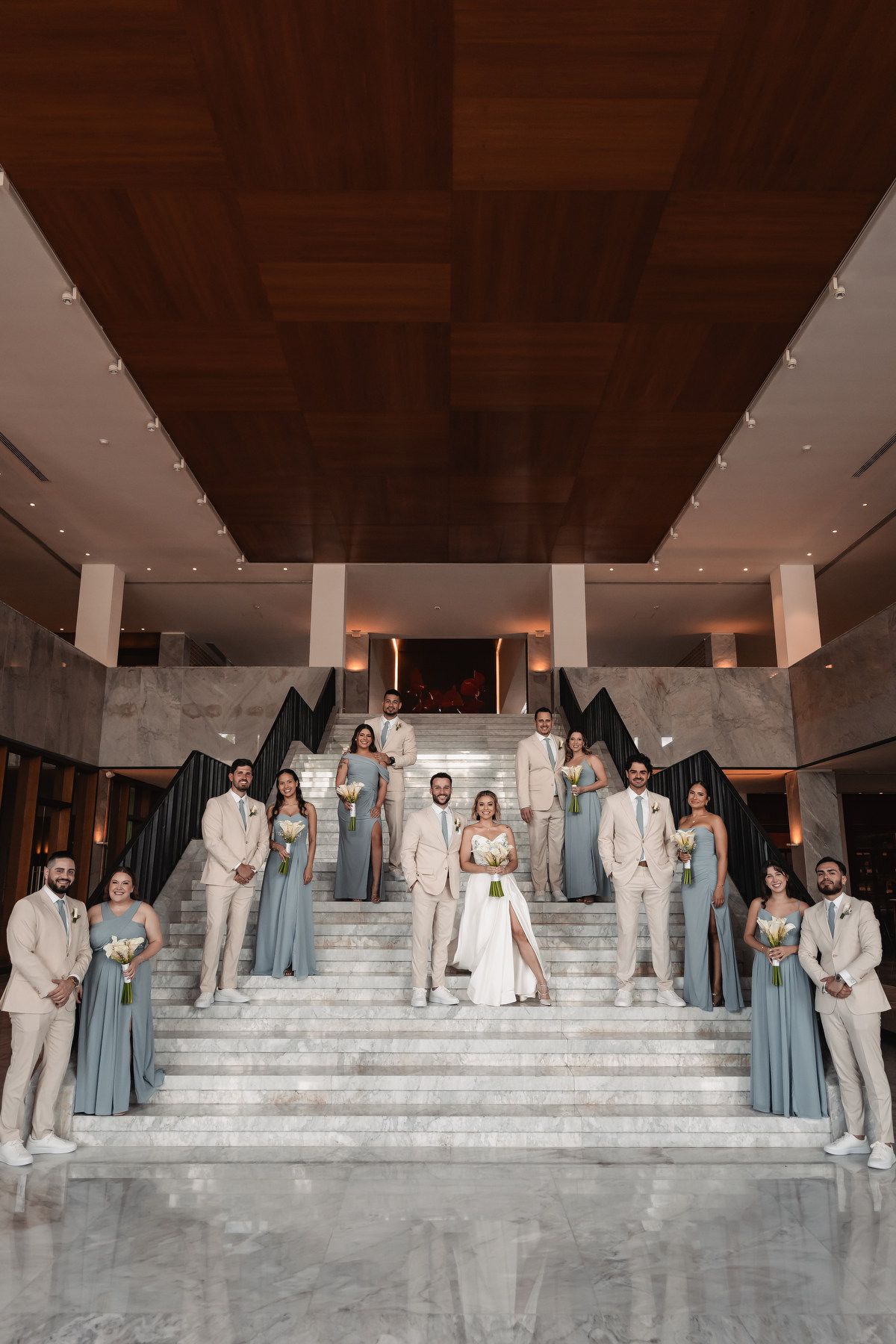 Wedding party poses on marble staircase in beige suits and dusty blue bridesmaid gowns.