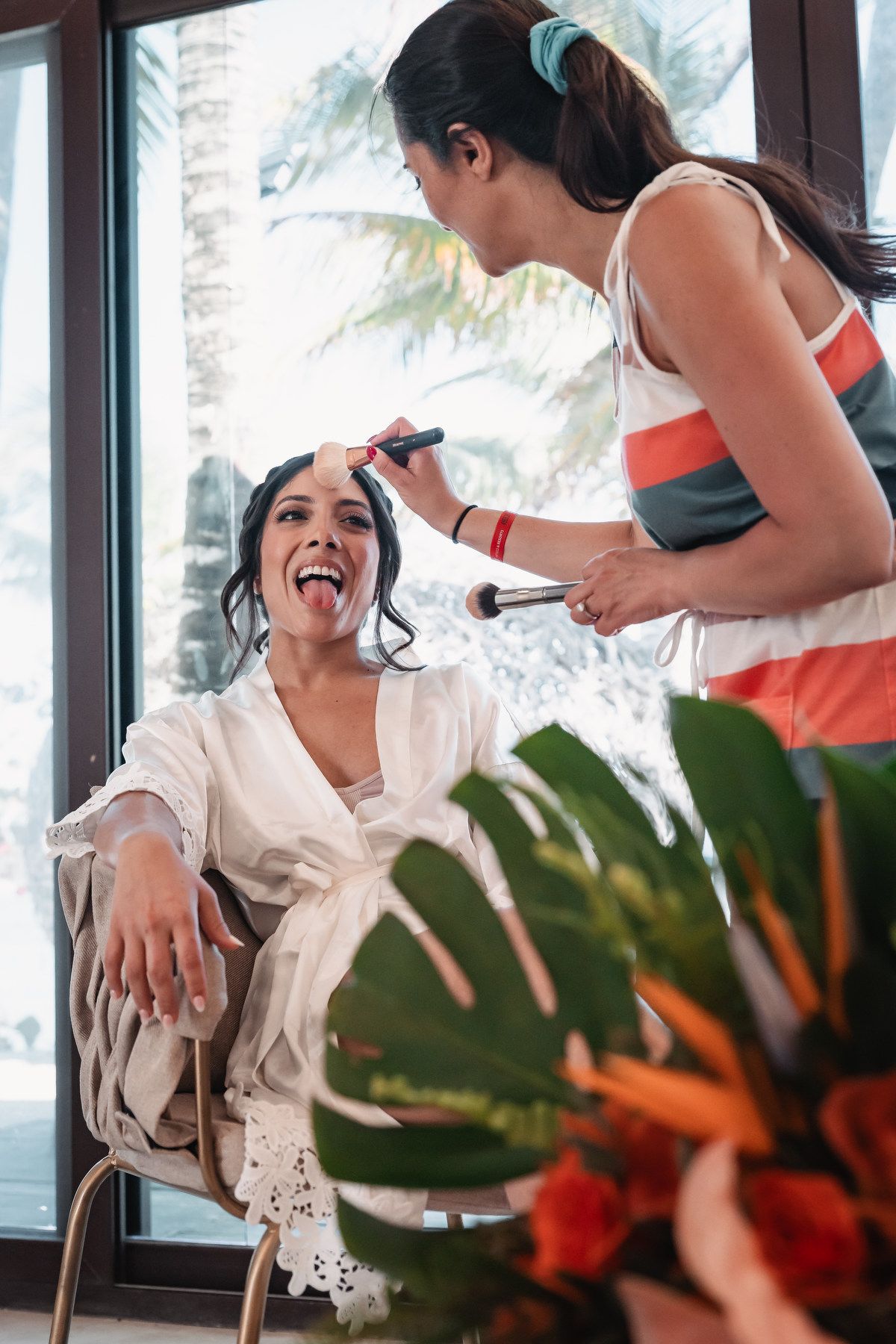 Bride laughing with tongue out while makeup artist applies makeup before beach wedding ceremony.