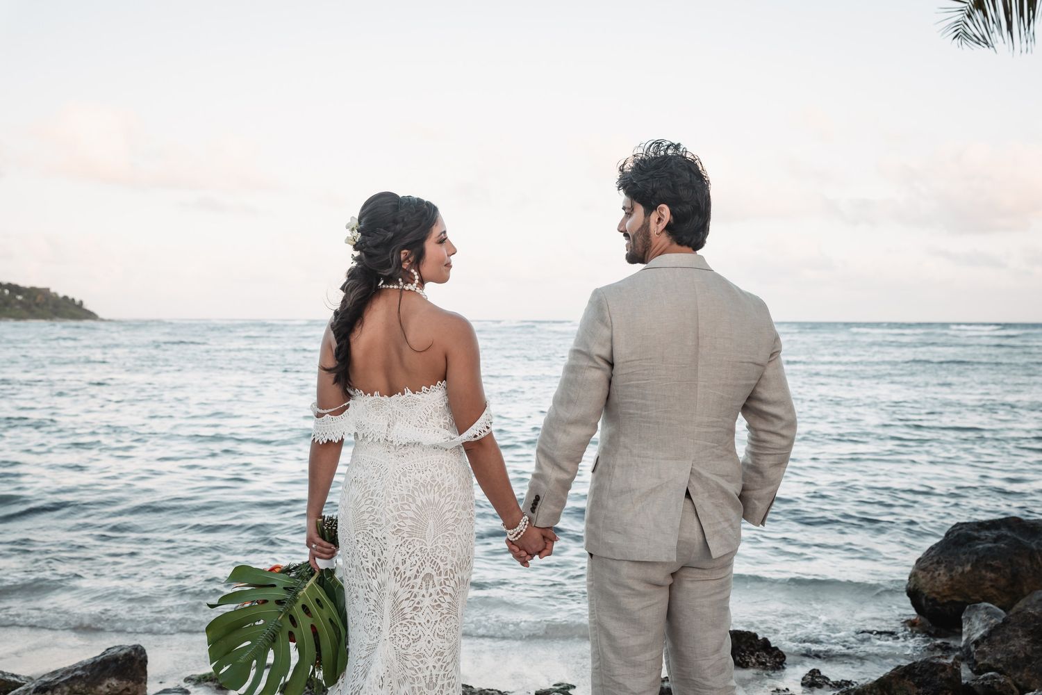 Bride and groom hold hands facing ocean at sunset on tropical beach wedding day.