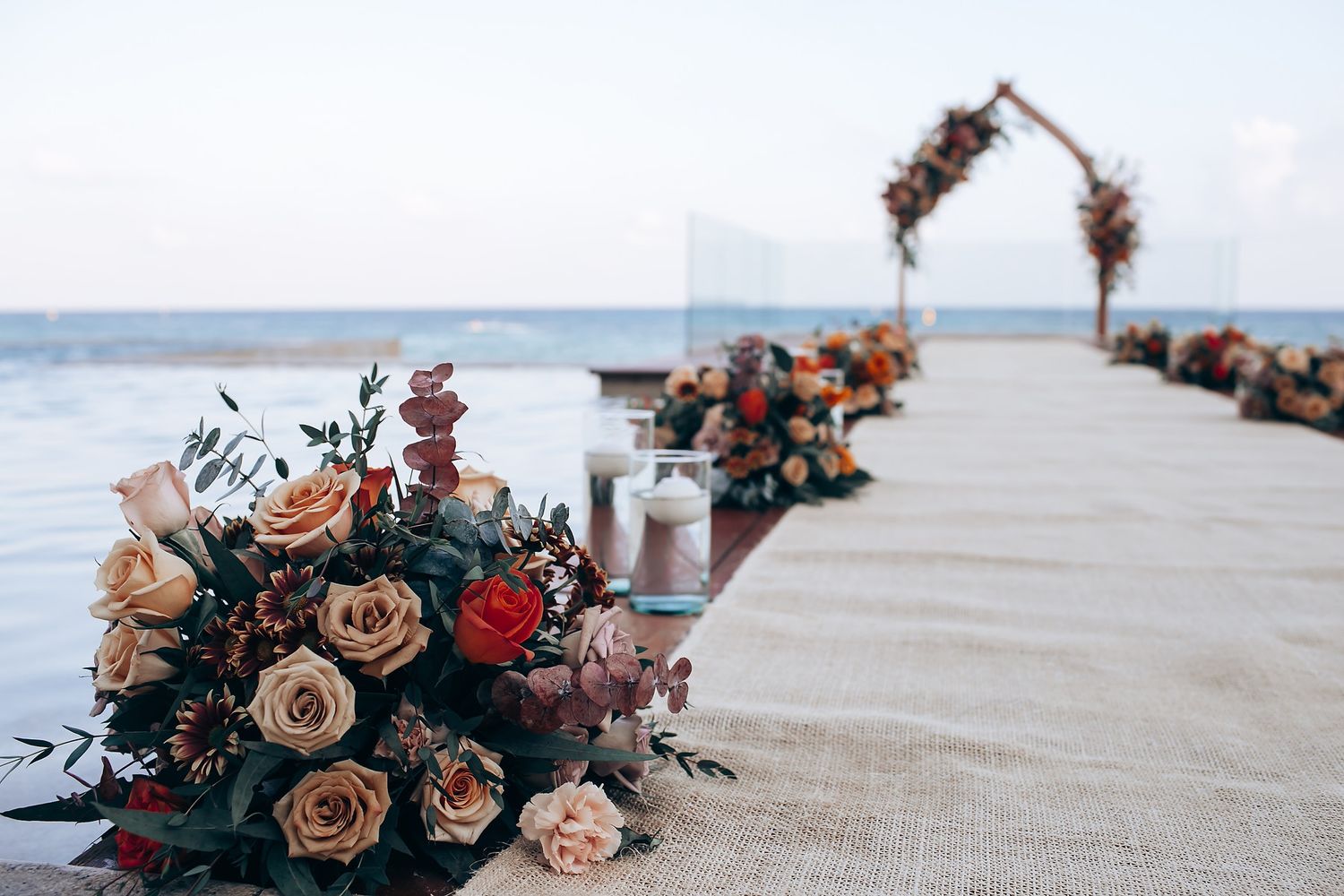 Beach wedding ceremony setup with floral arch and rose bouquets lining a wooden pier over ocean waters.