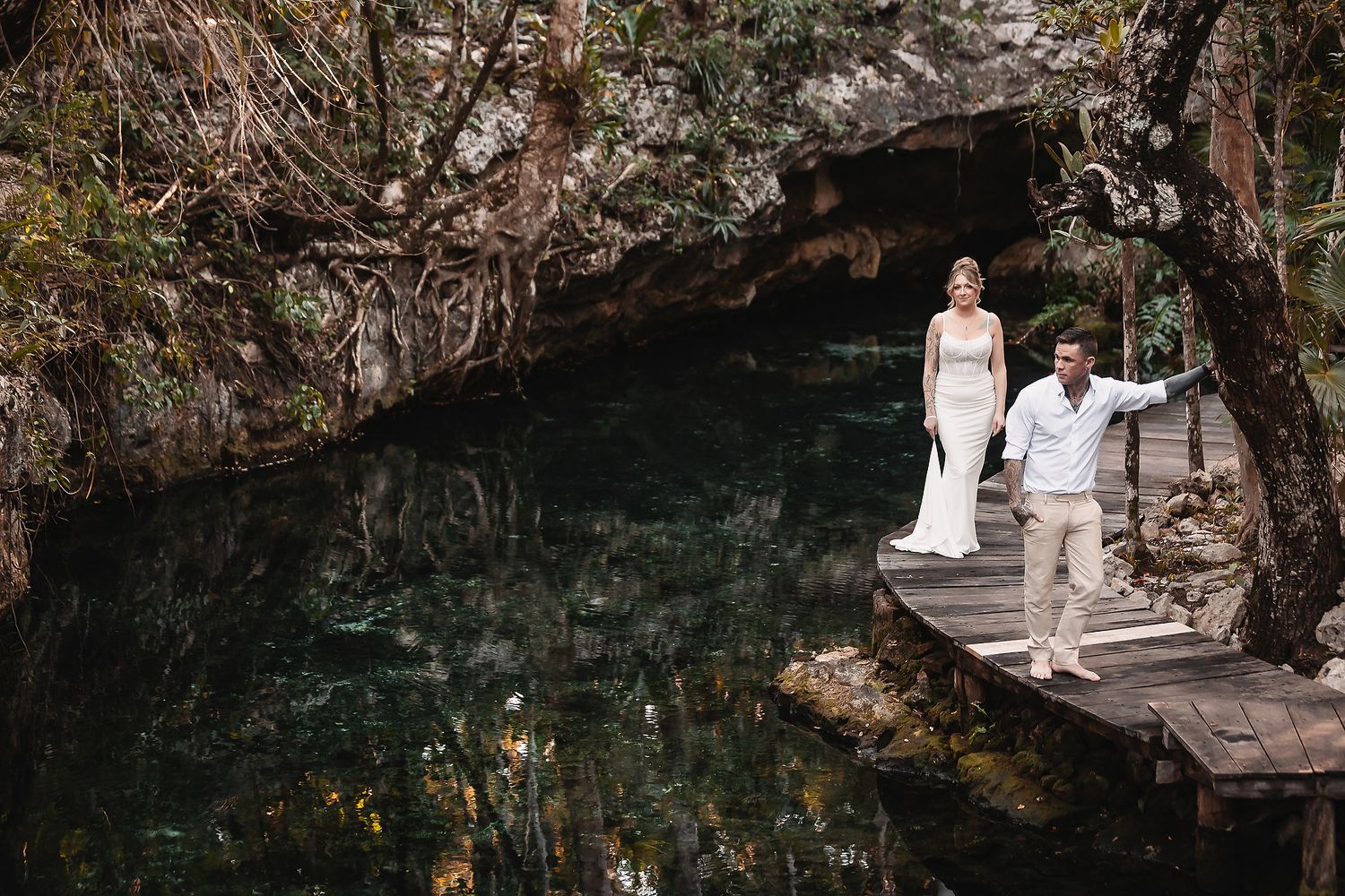Bride and groom walk hand-in-hand on a wooden dock over a serene cenote surrounded by lush tropical greenery.