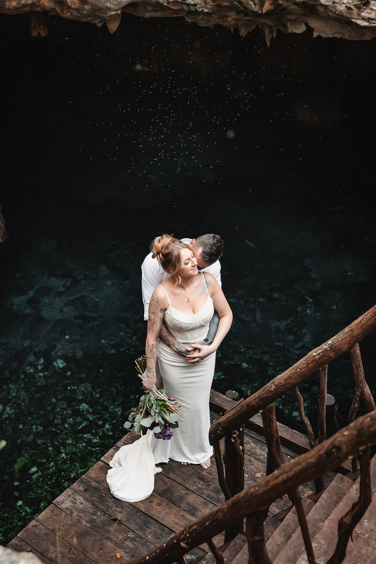 Bride and groom share a romantic moment on wooden steps above a turquoise cenote, bride holding a floral bouquet.